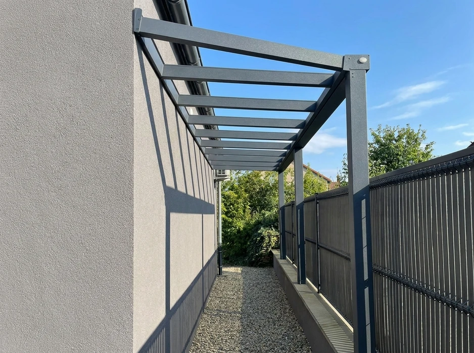 Pergola aluminium anthracite adossée à un mur de bâtiment, couvrant un passage étroit entre façade enduit gris et clôture rigide à lames verticales